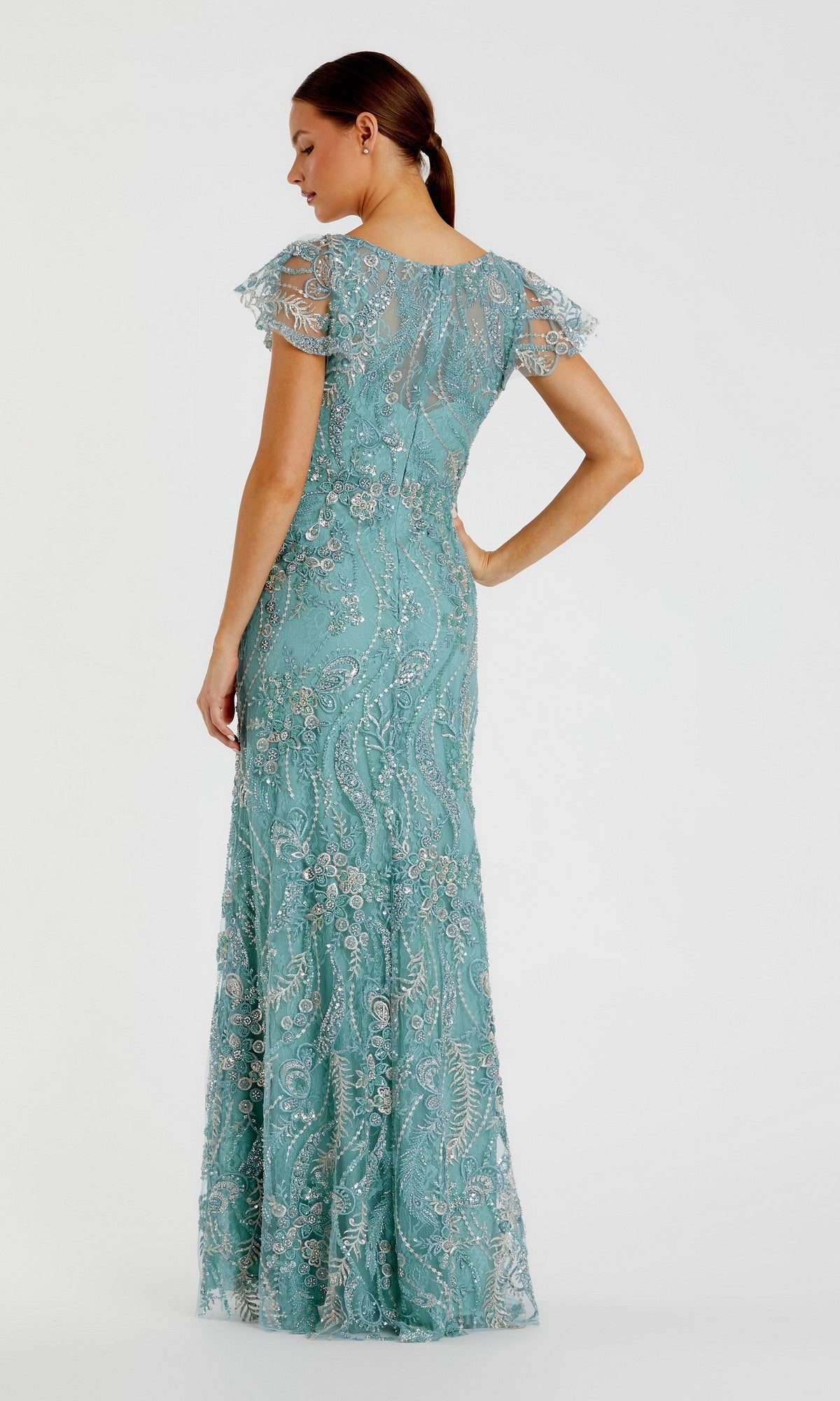 A woman stands against a plain background, one hand on her hip, wearing the Long Formal Dress: Mac Duggal 68914—a floor-length, light blue lace gown with short sleeves that exudes timeless elegance.